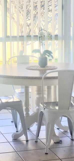 A white dining table with chairs, a potted plant, and a window with vertical blinds. - Glenmore Cottage (Glenmore Park)