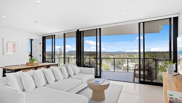 A modern living room with a white sofa, a round coffee table, and a view of a balcony with a cityscape and mountains in the background. - Relax in luxury, a short walk to the beach (Gold Coast)