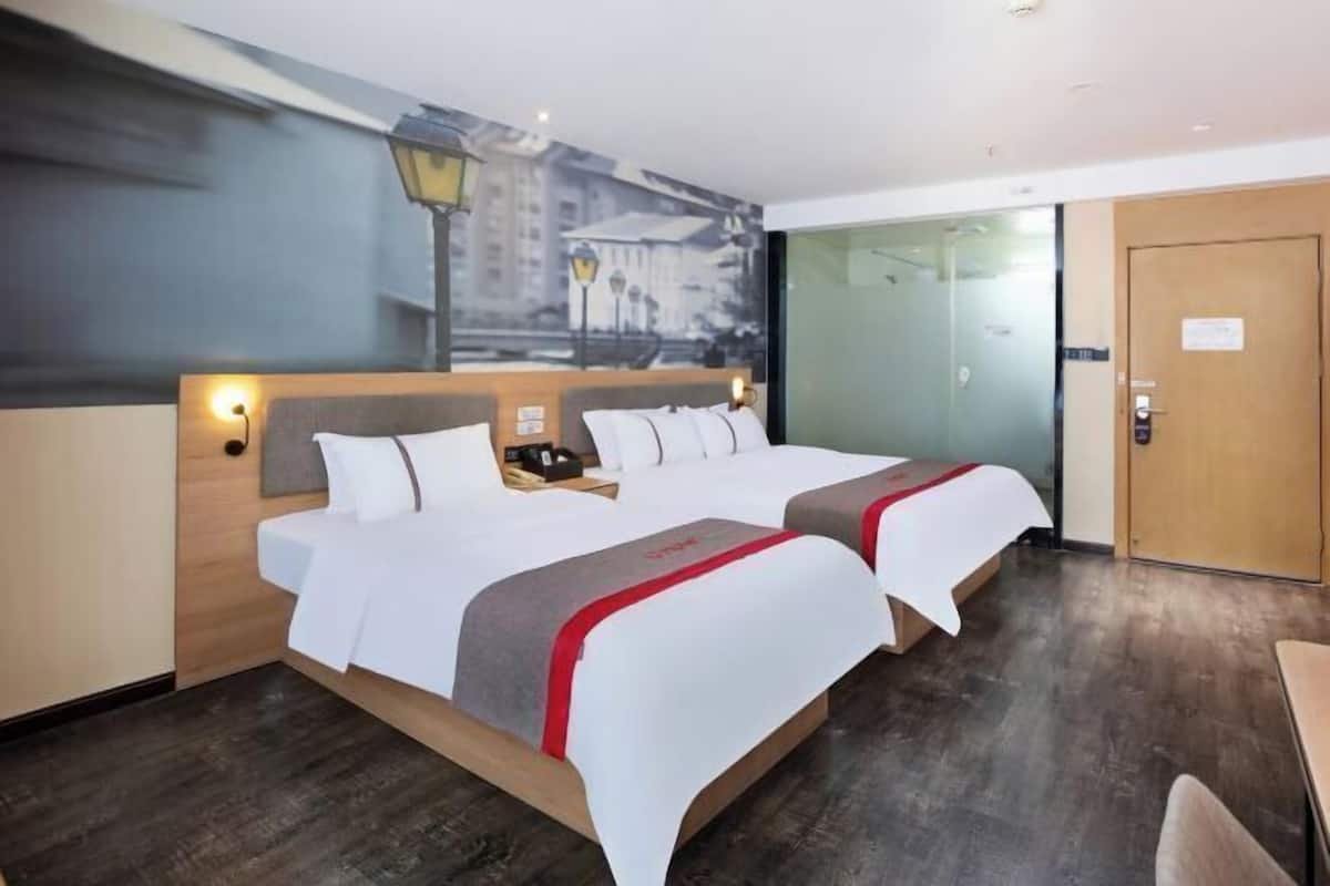 A hotel room with two beds, a wooden headboard, a nightstand, a lamp, and a cityscape mural on the wall.