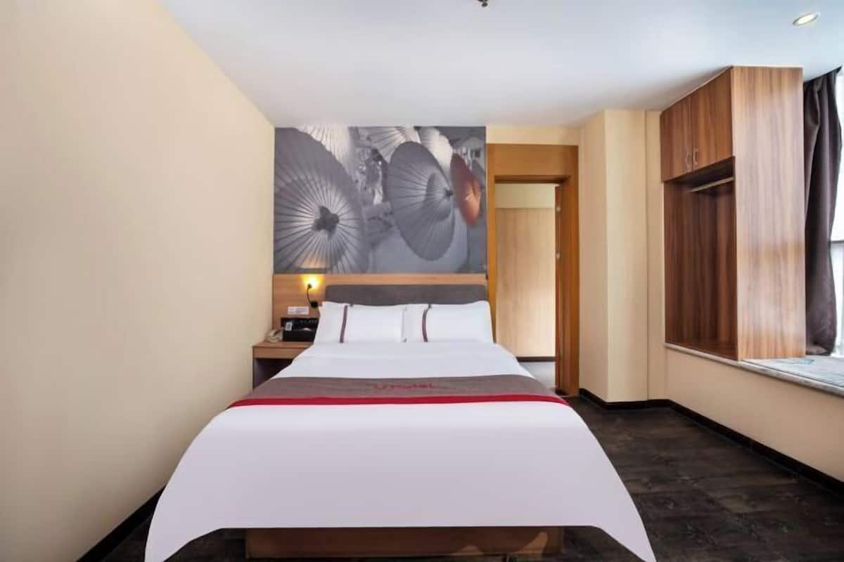 A hotel room with a large bed, a nightstand, a wall-mounted lamp, and a window with curtains.