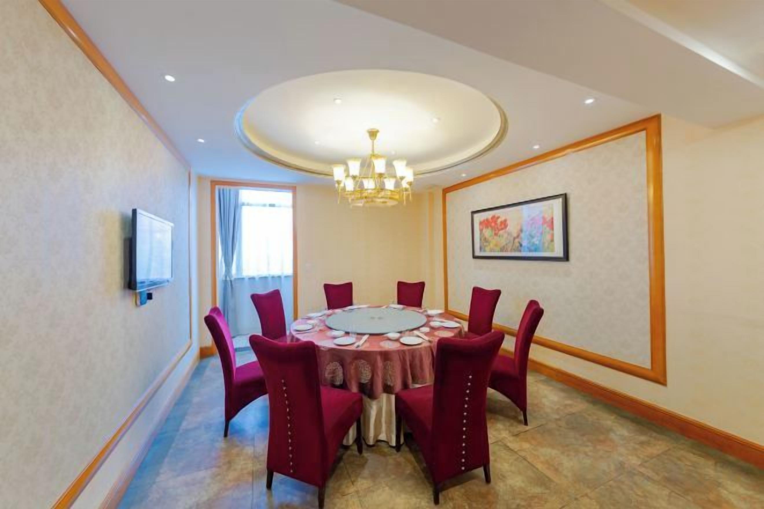 A conference room with a round table, red chairs, a chandelier, a television, and a painting on the wall.