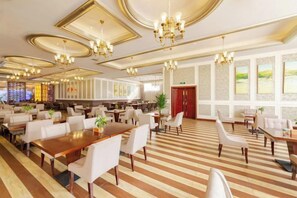 Un vaste espace de restauration d’hôtel, doté de nombreuses tables et chaises, de grandes fenêtres et d’éléments décoratifs au plafond.