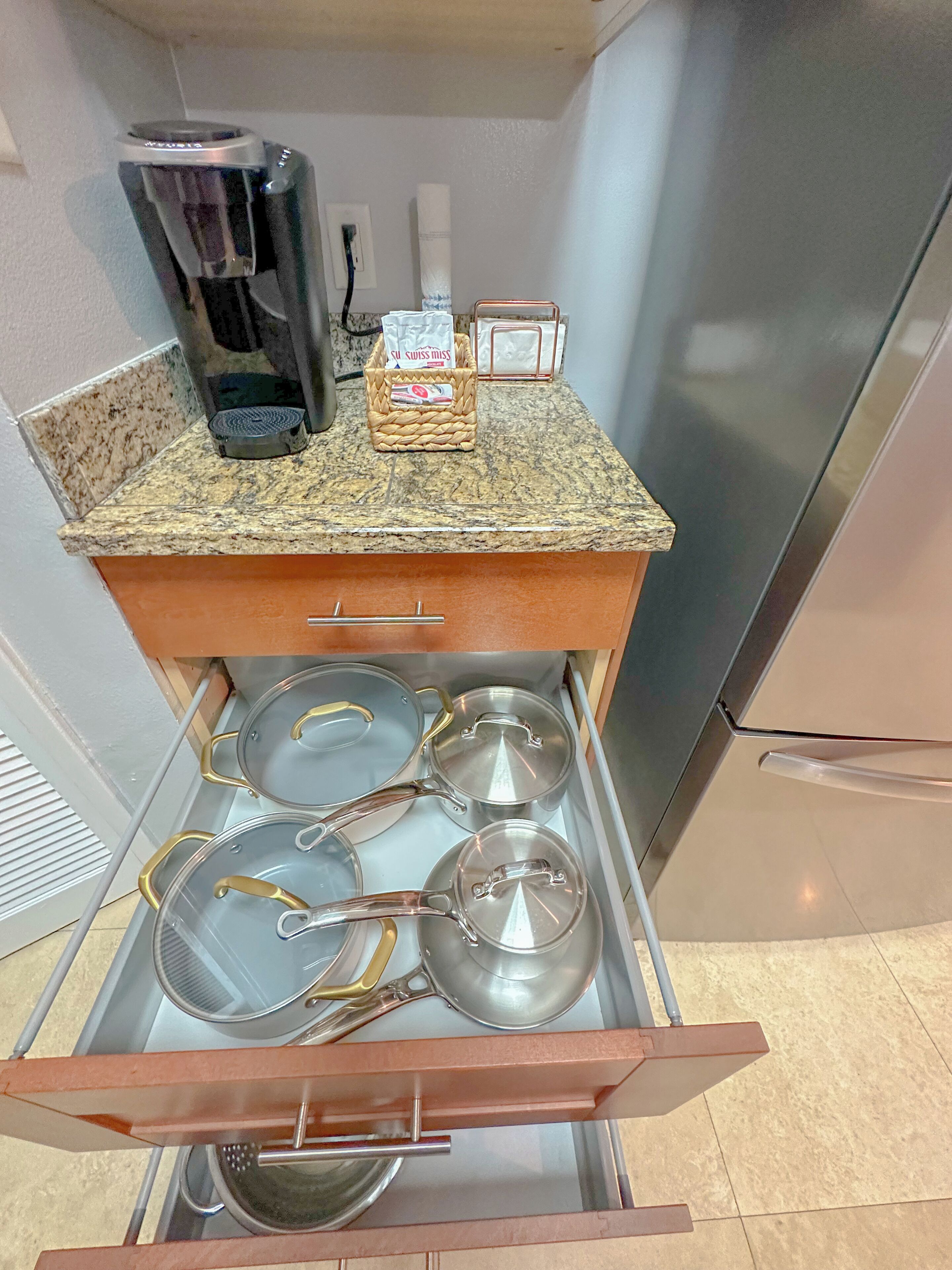 A kitchen with a drawer containing pots and pans, a coffee maker, and a basket with a Swiss Miss tea bag.
