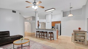 A modern living room with a dark sofa, a glass coffee table, a patterned rug, a kitchen area with a bar stool set, and a ceiling fan. - Charming 3-bedroom Pet Friendly house in tranquil Riverview (Riverview)