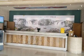 Un mostrador de recepción con un cuadro grande de una cascada y montañas, una pantalla de televisión y una pequeña mesa con una caja y una caja de pañuelos.