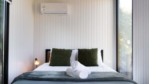 A neatly made bed with a grey blanket and two green pillows, an air conditioning unit on the wall, and a bedside lamp. - Tussock Views by Tiny Away (Burkes Pass)