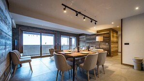 Un espace repas moderne avec une table et des chaises en bois, de grandes fenêtres et une vue sur l’extérieur.