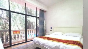 Une chambre avec une grande fenêtre, un lit recouvert de draps blancs et d’une couverture rouge et or, et un plancher en bois.