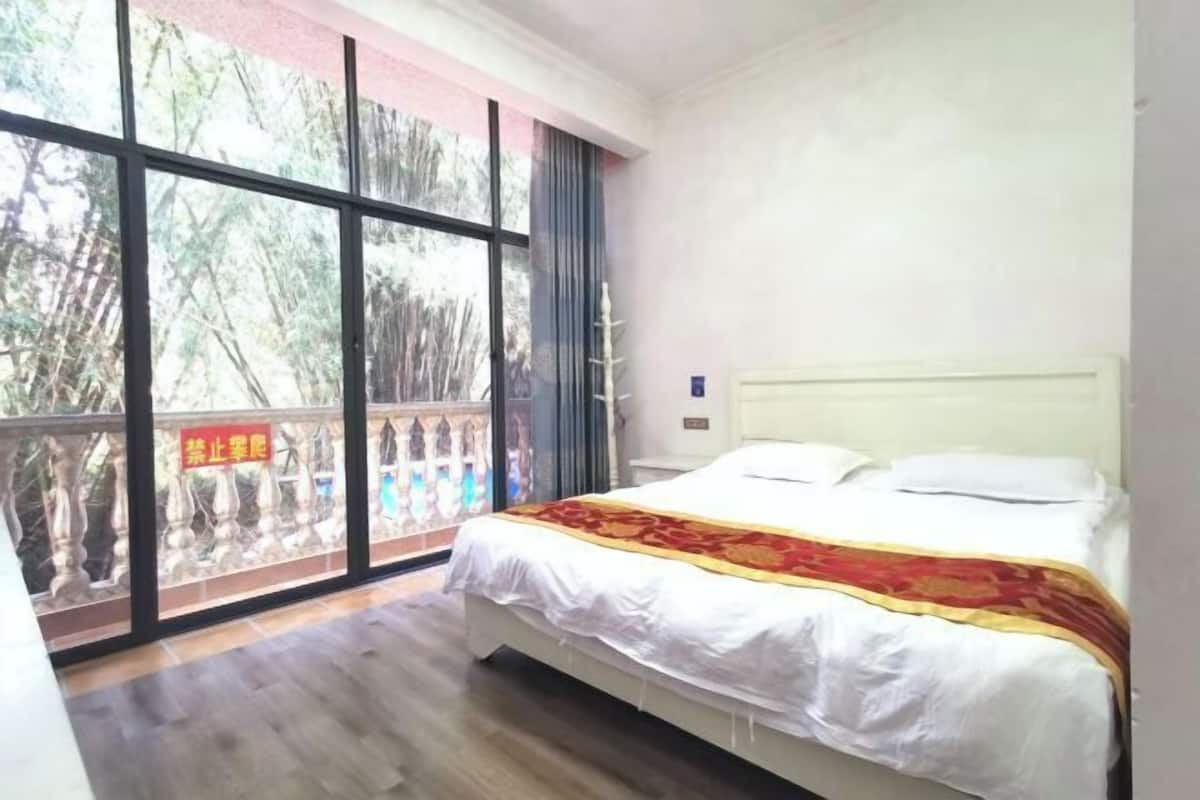 A bedroom with a large window, a bed with white bedding and a red and gold blanket, and a wooden floor.