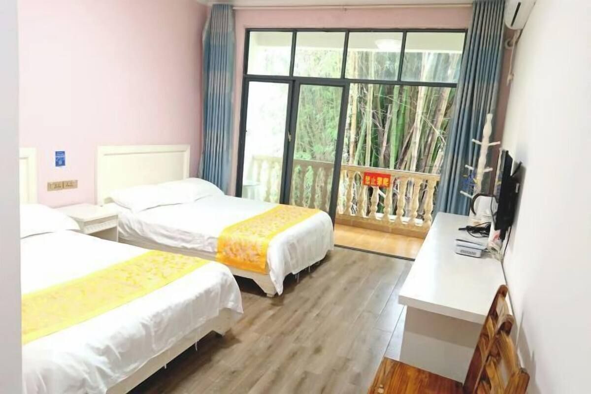 A hotel room with two beds, a desk, a window with a view of trees, and a balcony with a railing.