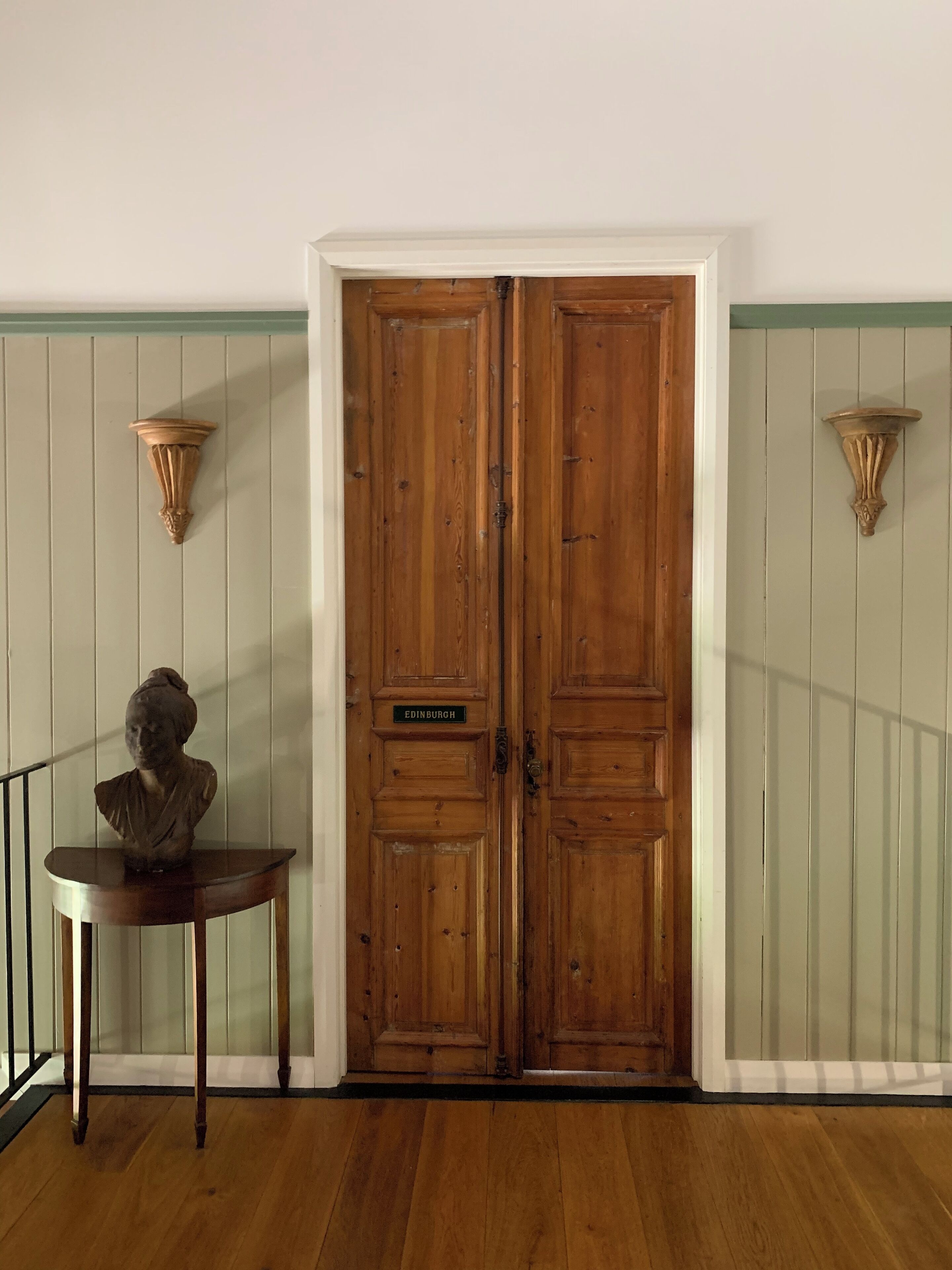 A wooden double door with a plaque reading "EDINBURGH," a bust on a small table, and two wall-mounted sconces.