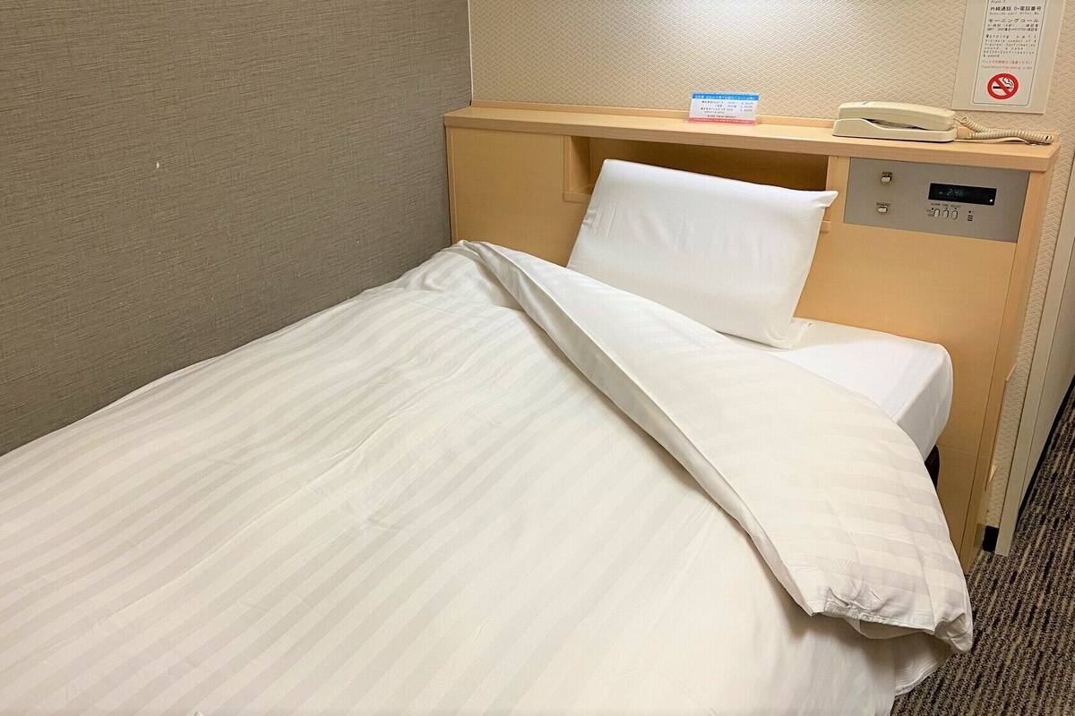 Un lit simple avec une literie blanche, une tête de lit en bois, une table de chevet avec un téléphone et une petite carte, et un panneau de commande fixé au mur.