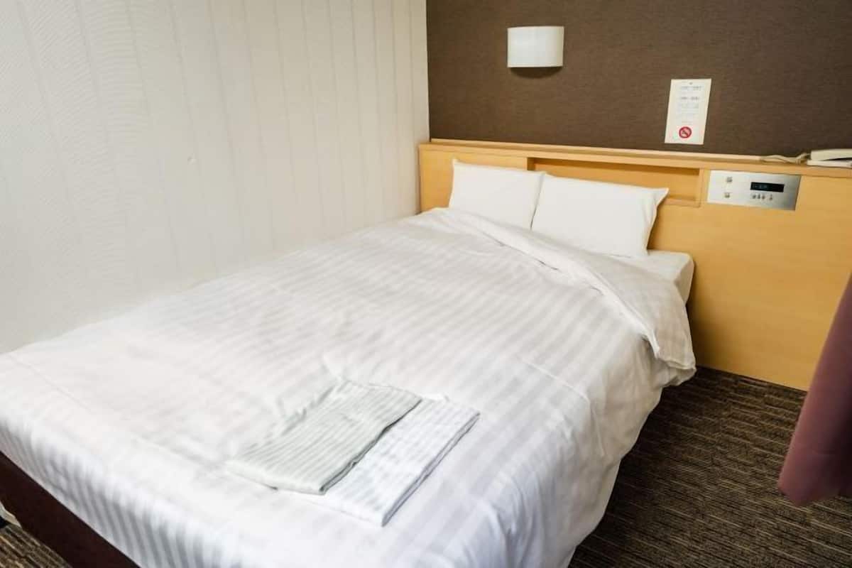 Une chambre d’hôtel équipée d’un lit, d’une lampe fixée au mur, d’un panneau de commande et d’une fenêtre avec des stores.
