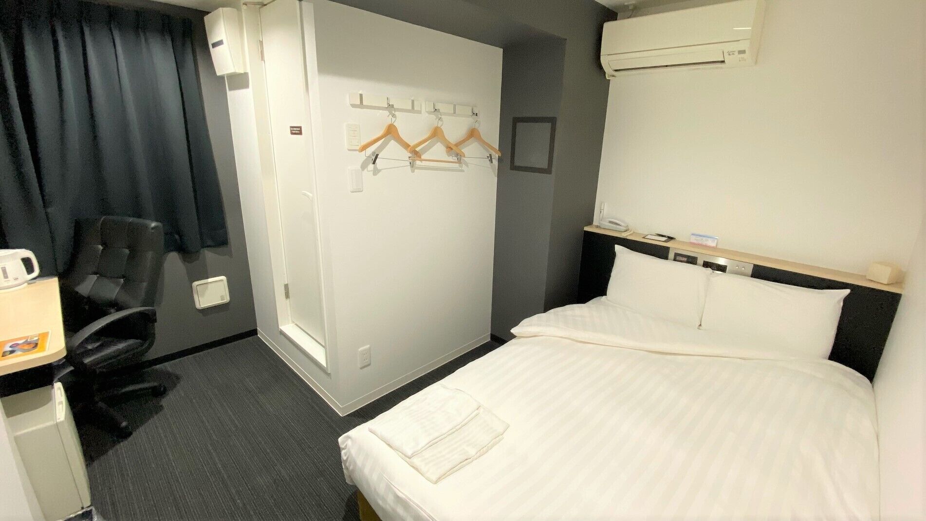Ein Hotelzimmer mit Bett, Schreibtisch mit Stuhl, Kleiderhaken und einer Wand-Klimaanlage.