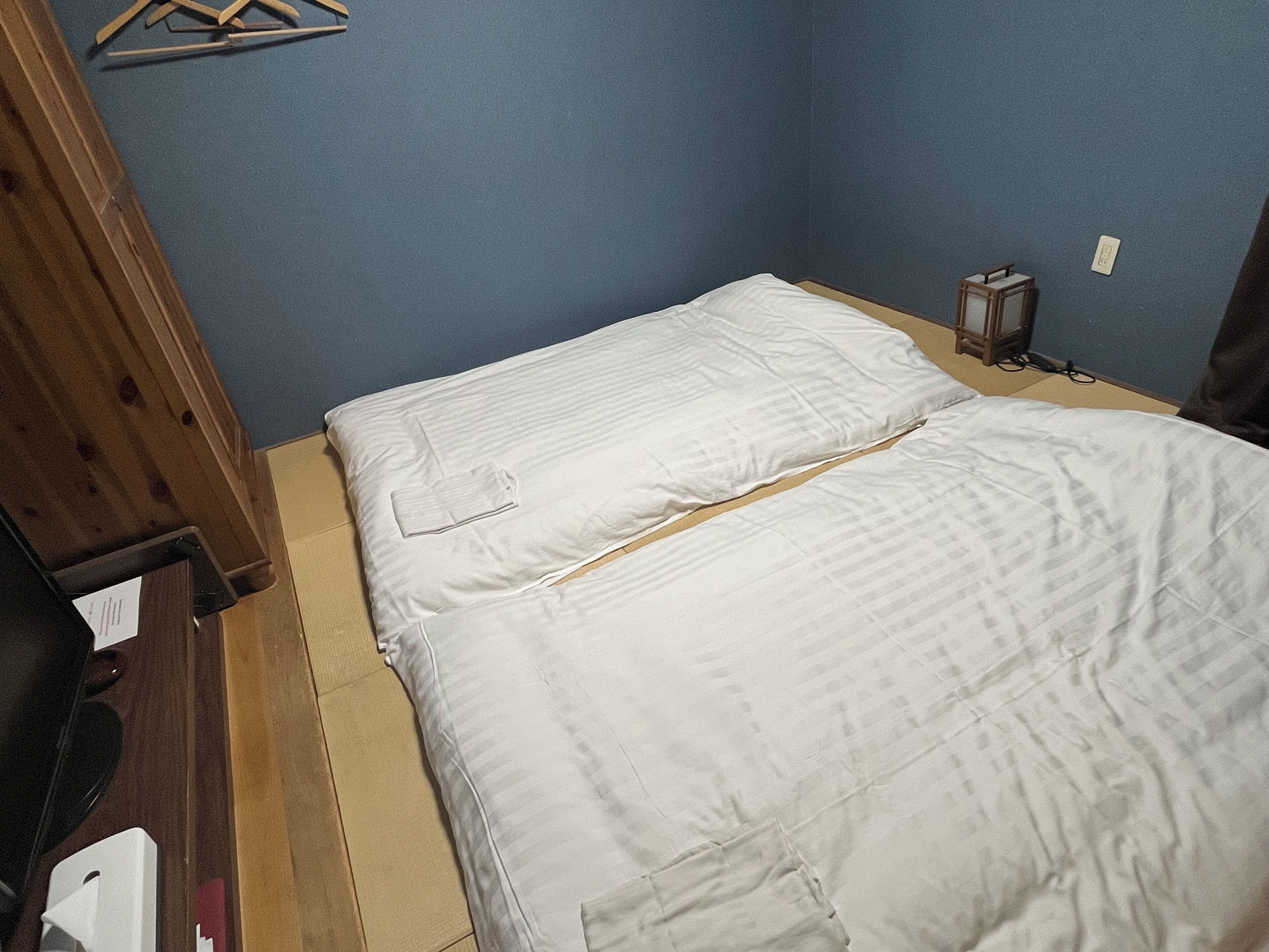 Una habitació petita amb un llit, una petita arca de fusta i una làmpada fixada a la paret.