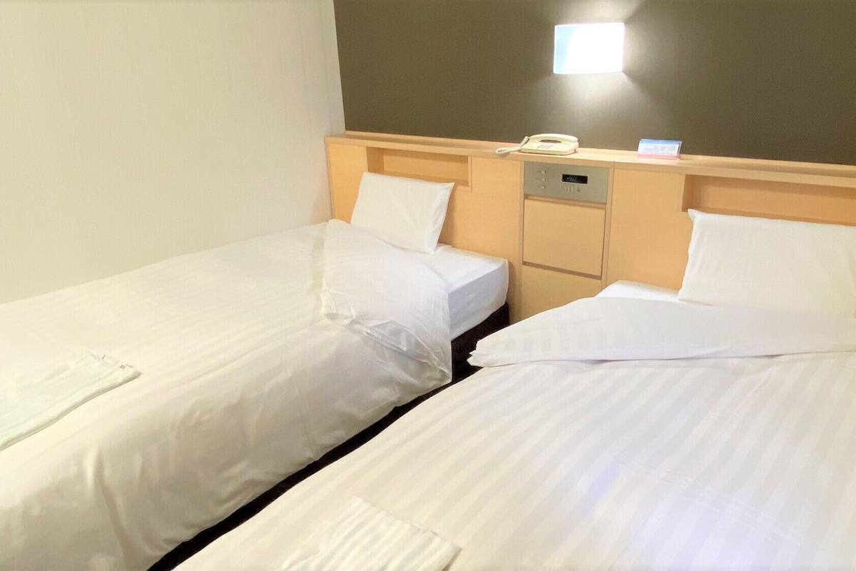 Deux lits simples avec du linge de lit blanc, une tête de lit en bois, une table de chevet avec un téléphone et une lampe fixée au mur.