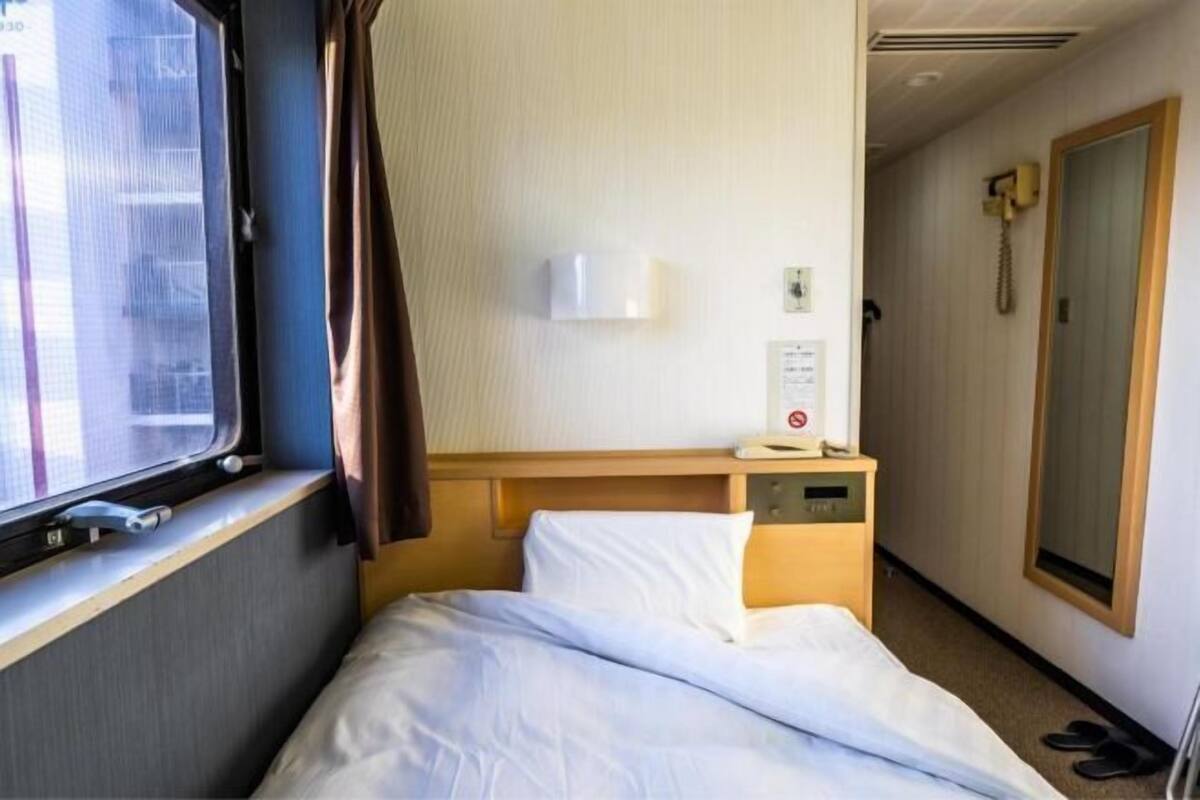 Un lit simple avec une literie blanche, une petite table de chevet avec une lampe, une fenêtre avec des rideaux et une porte avec un téléphone.