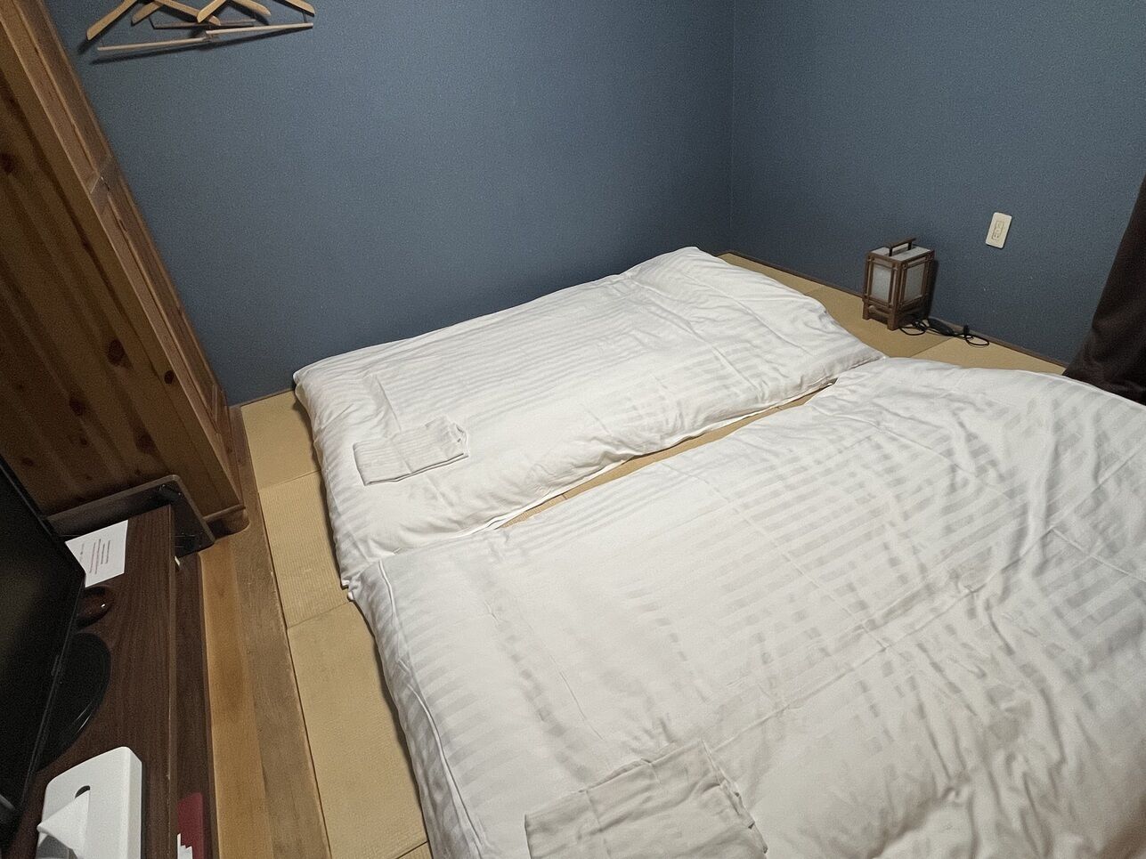 Ein kleines Zimmer mit einem Einzelbett, einem Holzschrank, einem kleinen Tisch und einer Wandleuchte.