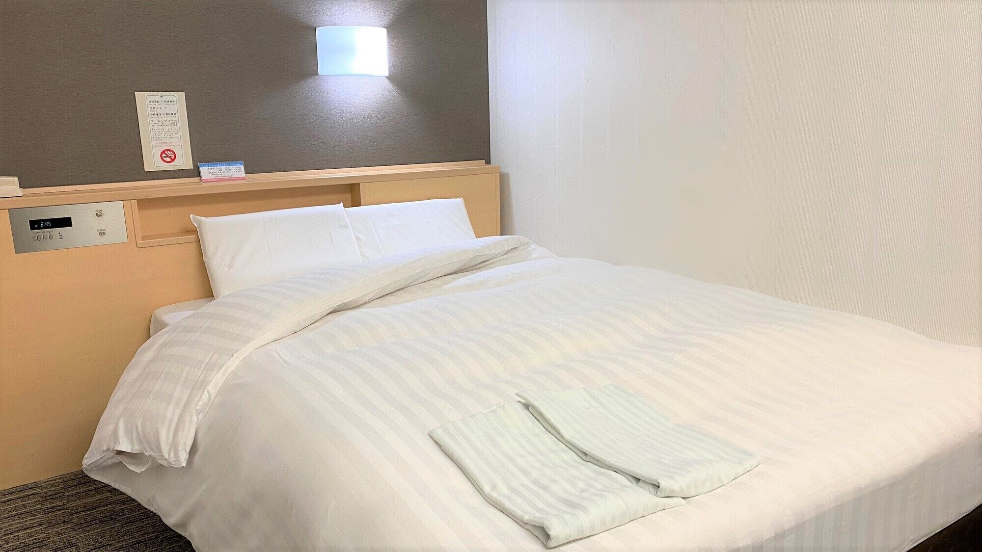 Habitació d'hotel amb un llit, un panell de control al costat del llit, una làmpada penjada a la paret i un cartell amb text.