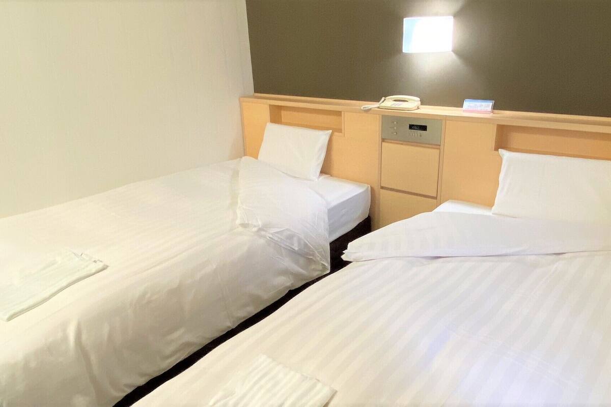 Deux lits simples avec du linge de lit blanc, une tête de lit en bois, une table de chevet avec un téléphone et une petite horloge numérique.