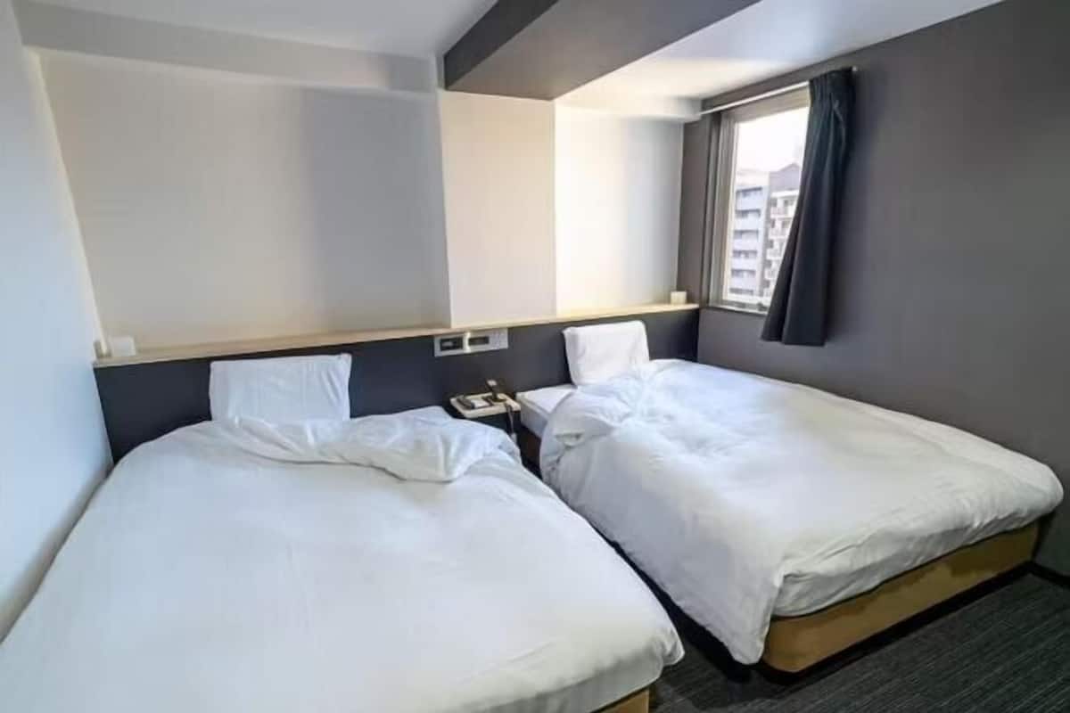 Une chambre d’hôtel avec deux lits simples, une table de chevet avec un téléphone, une fenêtre avec des rideaux et un luminaire fixé au mur.