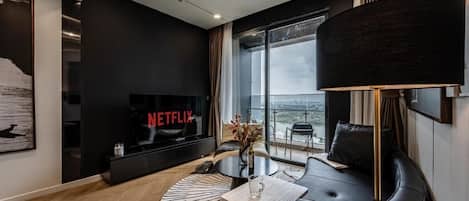 A modern hotel room with a large window, a flat-screen TV, a round coffee table, and a sofa.
