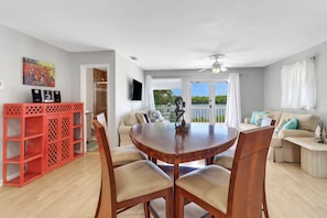 Un vaste espace de vie comprenant une table à manger ronde en bois, des chaises rembourrées de couleur beige, une étagère rouge, un canapé beige agrémenté de coussins turquoise et une vue sur l’extérieur grâce à de grandes fenêtres.