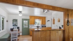 A kitchen with wooden cabinets, a wooden island, and a dining area with a white chair. - Central Heat & Air, WiFi, Wall Mural and Fully fenced minutes from downtown (Idyllwild-Pine Cove)