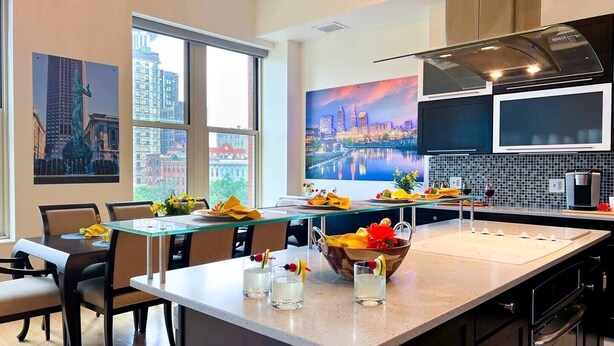 A modern kitchen with a large island, a dining area with a glass table, and artwork depicting cityscapes. - Downtown Cleveland: Steps To Sports & Events 2 Bedroom Condo by RedAwning (Cleveland)
