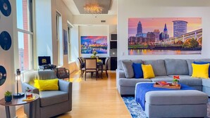 A modern living room with a grey sofa, yellow cushions, a dining area with a table and chairs, and a large cityscape painting on the wall. - Downtown Cleveland: Steps To Sports & Events 2 Bedroom Condo by RedAwning (Cleveland)