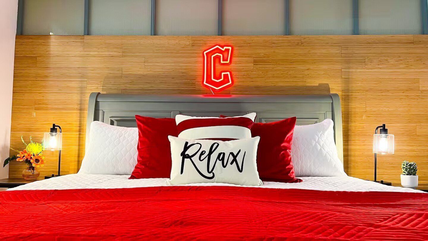 Ein ordentlich bezogenes Bett mit roter Bettwäsche und einem Kissen mit der Aufschrift „Relax“, ein hölzernes Kopfteil und ein leuchtendes 'C'-Schild darüber.