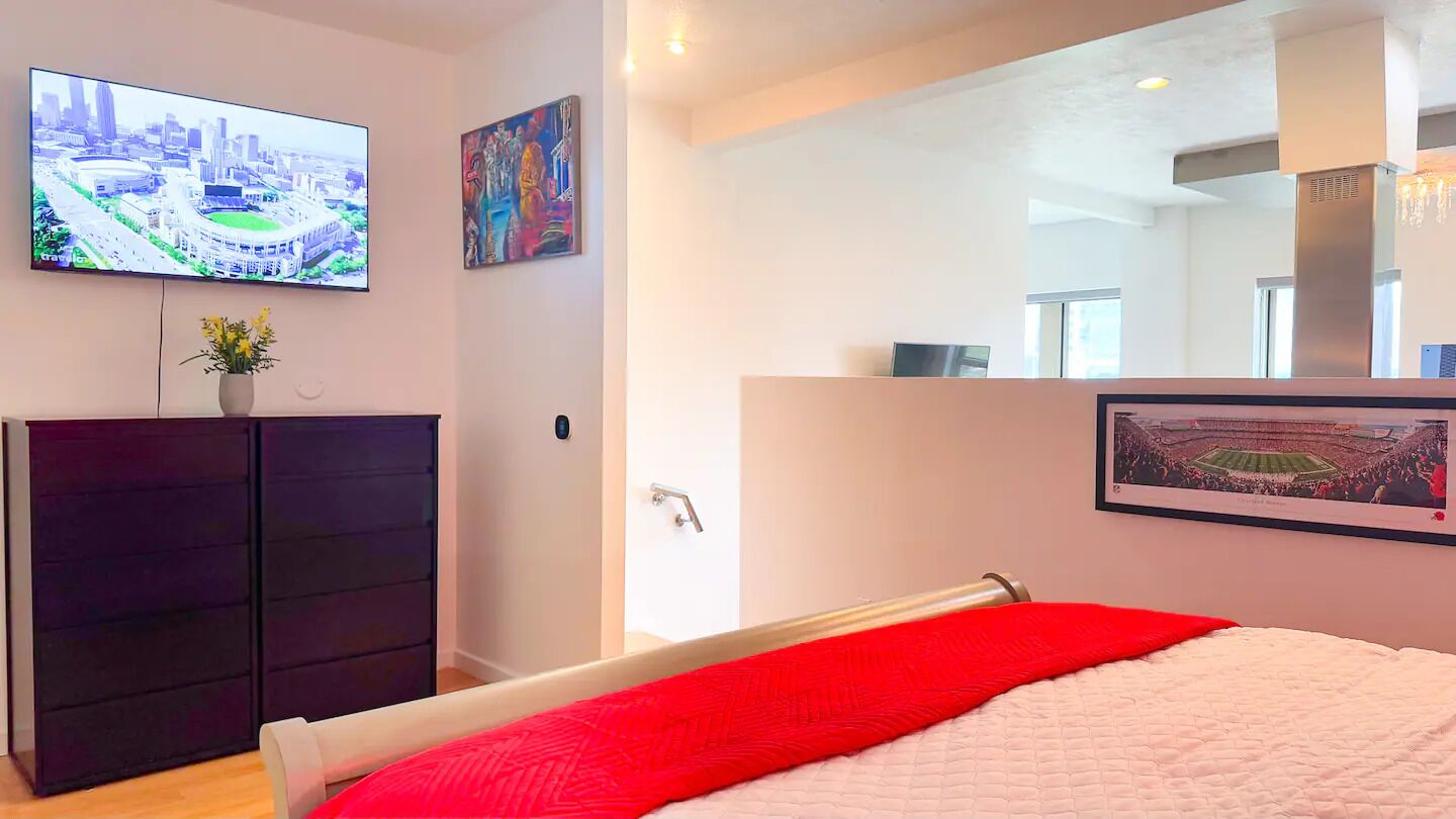 A bedroom with a bed, a TV mounted on the wall, a painting, and a mirror reflecting the room.