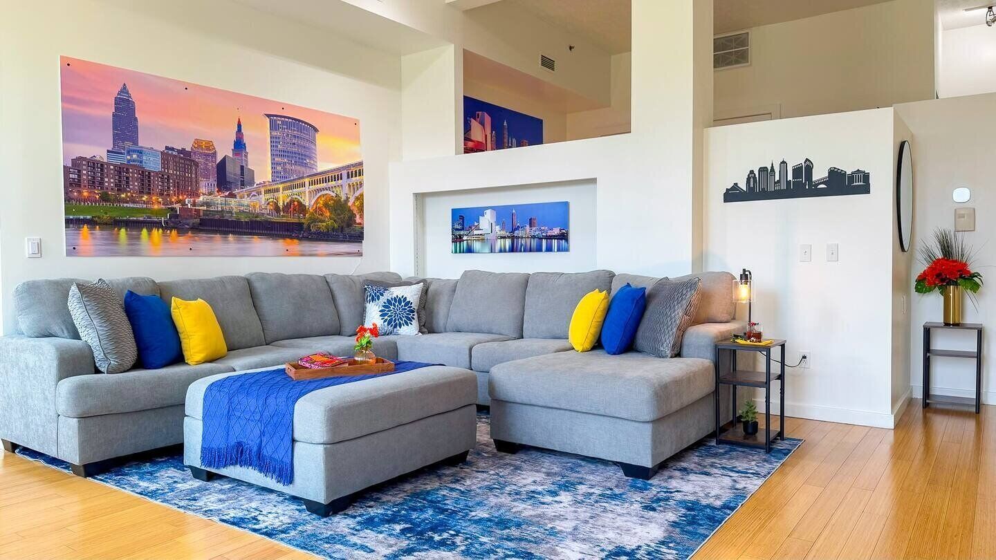 A modern living room with a sectional sofa, a coffee table, and a rug. There are framed cityscape paintings on the walls.