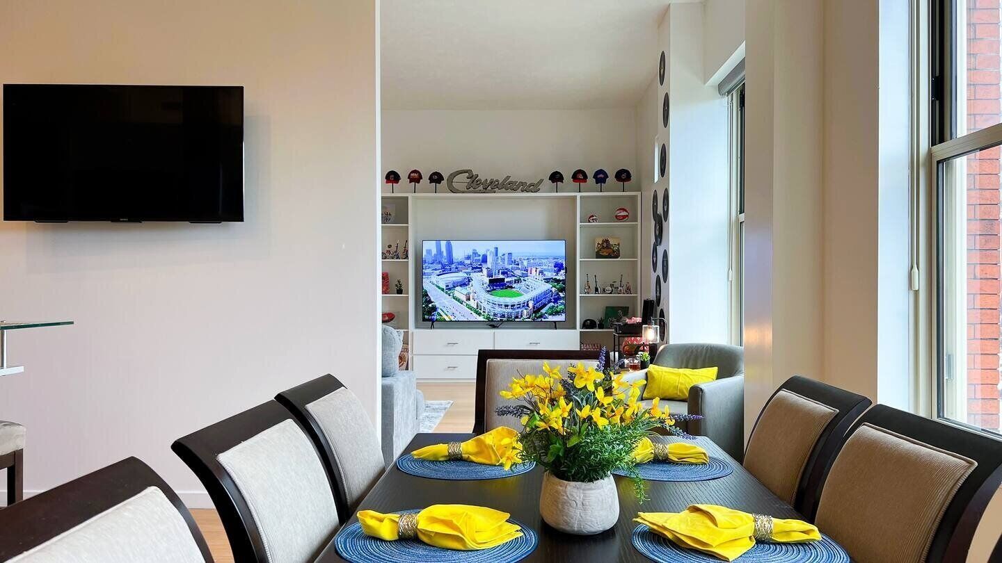 A dining area with a table set for four, a flat-screen TV in the background, and a shelf with hats above it.