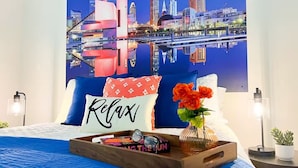 A hotel room with a cityscape view, a bed with a blue bedspread, a tray with books and a vase of flowers, and a pillow with the word "Relax". - Downtown Cleveland: Steps To Sports & Events 2 Bedroom Condo by RedAwning (Cleveland)