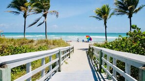 A beach pathway with wooden railings, palm trees, and a colorful umbrella on the sand.