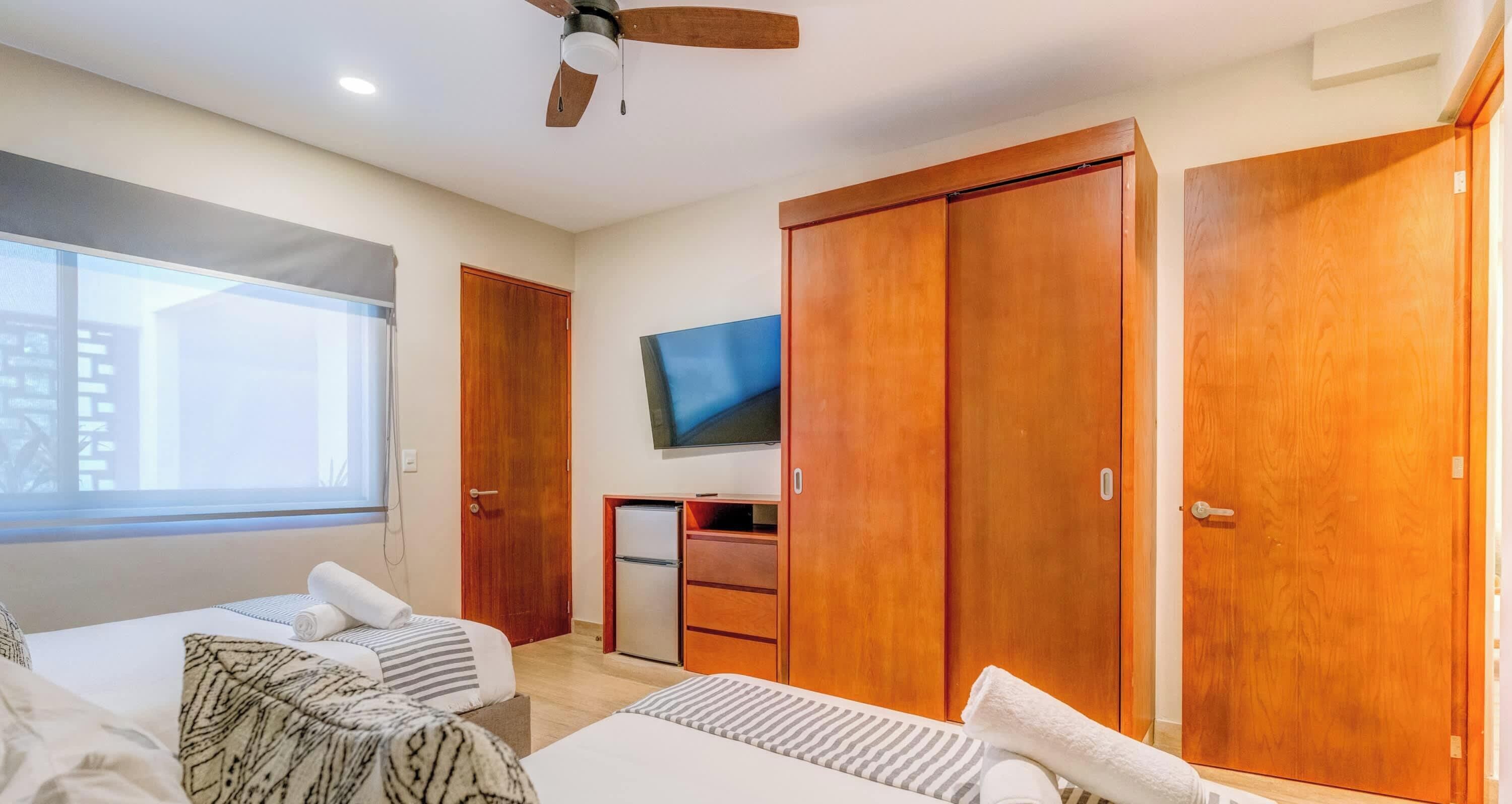 Une chambre d’hôtel avec deux lits, une télévision, un ventilateur de plafond, des armoires en bois et une fenêtre.