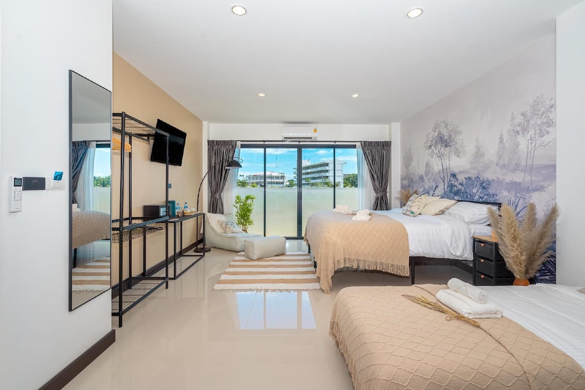 A modern hotel room with a large bed, a desk, a chair, a flat-screen TV, and a large window with a view of buildings and greenery.