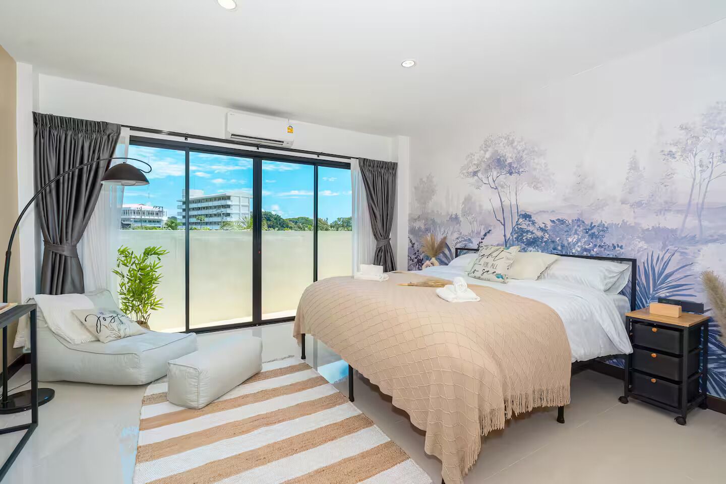 A modern bedroom with a large bed, a side table, a chair, and a view of the outdoors through a sliding door.
