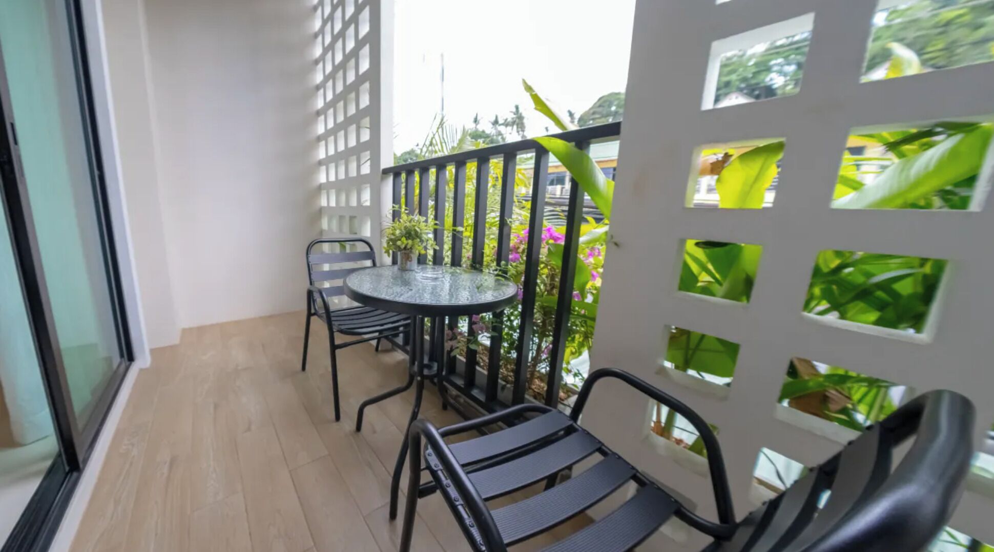 Ein Balkon mit rundem Glastisch und zwei Stühlen, umgeben von einem weißen Gitterzaun, durch dessen Öffnungen grüne Bepflanzung sichtbar ist.