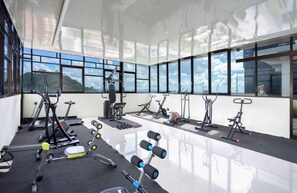 A gym with various exercise equipment, large windows, and a view of the outdoors. - Grand Valonia (Sumbersari)