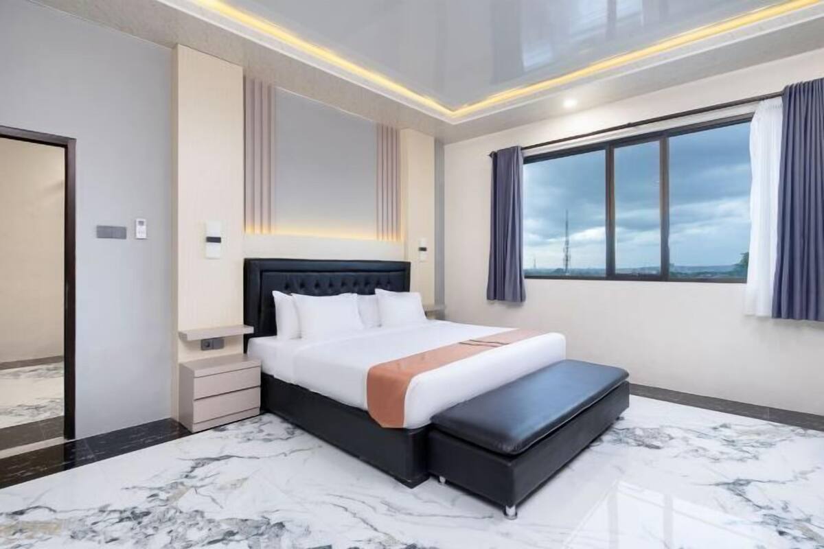 Une chambre d’hôtel moderne avec un grand lit, une banquette, une table de chevet et une grande fenêtre avec des rideaux.