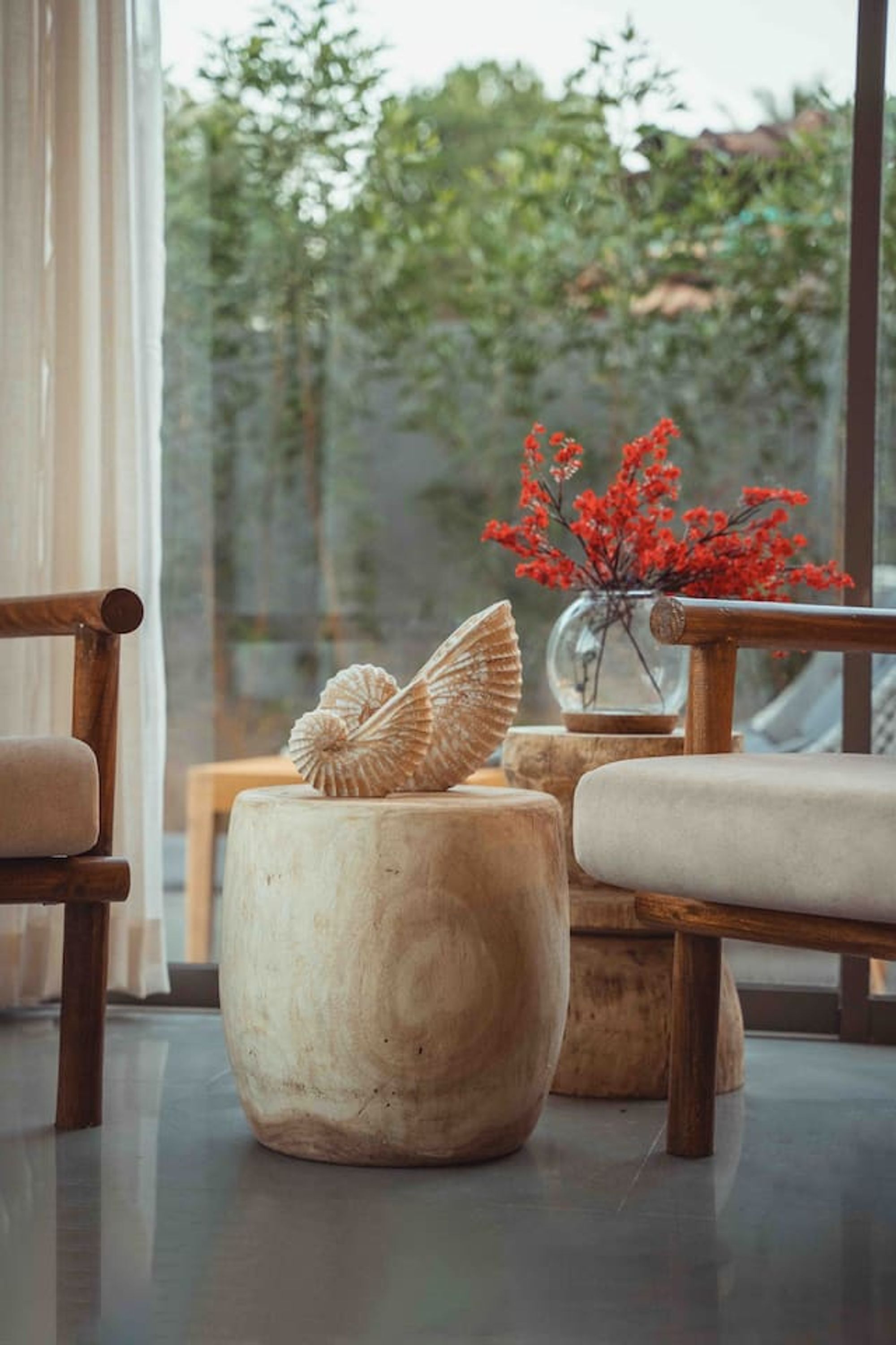 A wooden chair with a cushion, a round wooden stool, and a vase with red flowers on a windowsill.