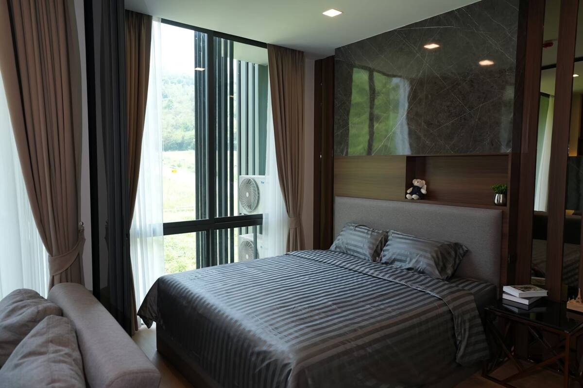 A modern hotel room with a large bed, a sofa, a desk with a chair, and a view of greenery outside.