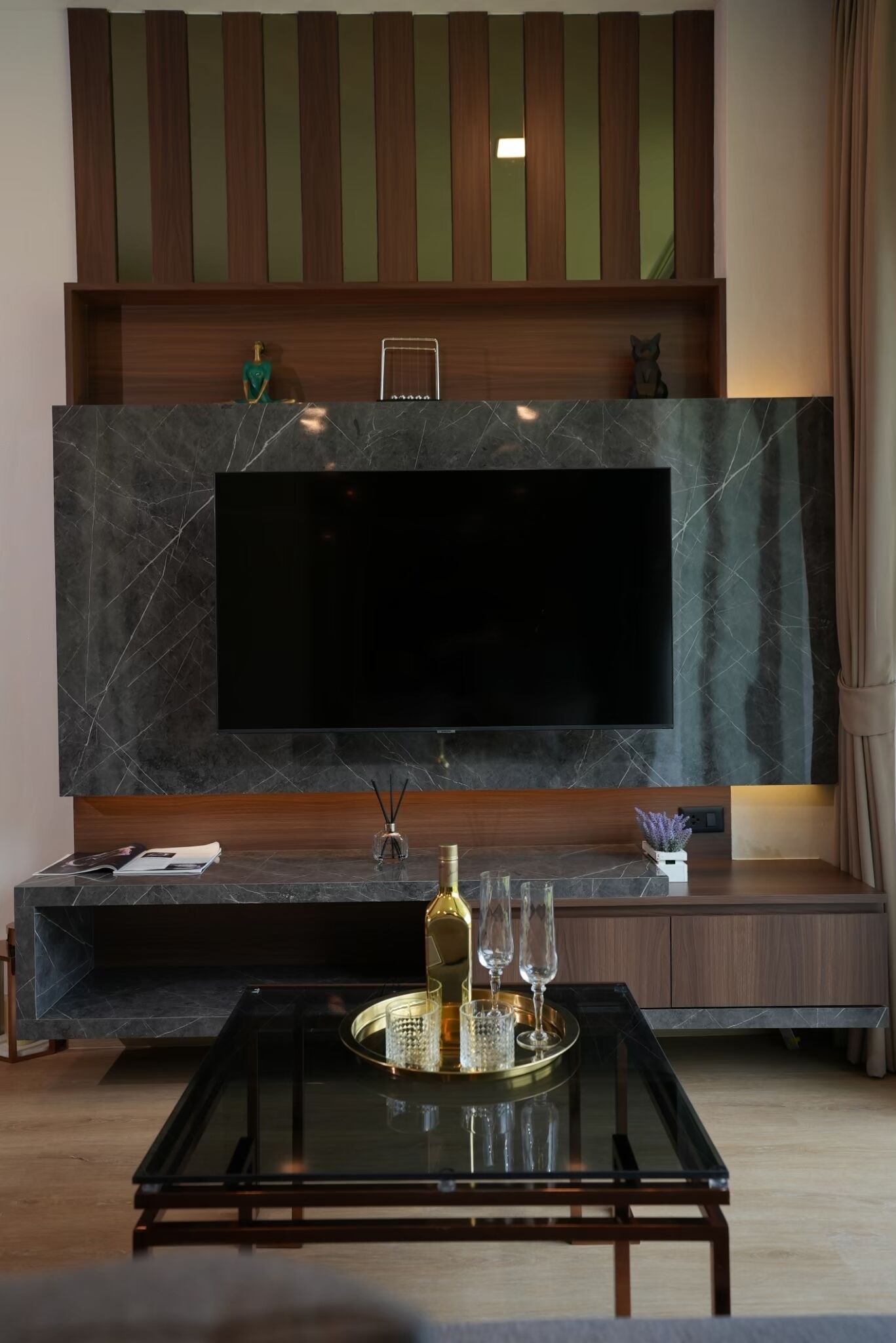 A modern living room with a large flat-screen TV mounted on a marble wall, a glass coffee table with decorative items, and a wooden entertainment unit above.