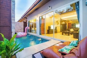 Una casa moderna con piscina, un flotador de flamenco rosa y una puerta de cristal que da a una sala de estar con sofá y una mesa de centro.