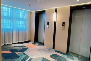 A hotel hallway with an elevator, a patterned carpet, and a window with curtains.