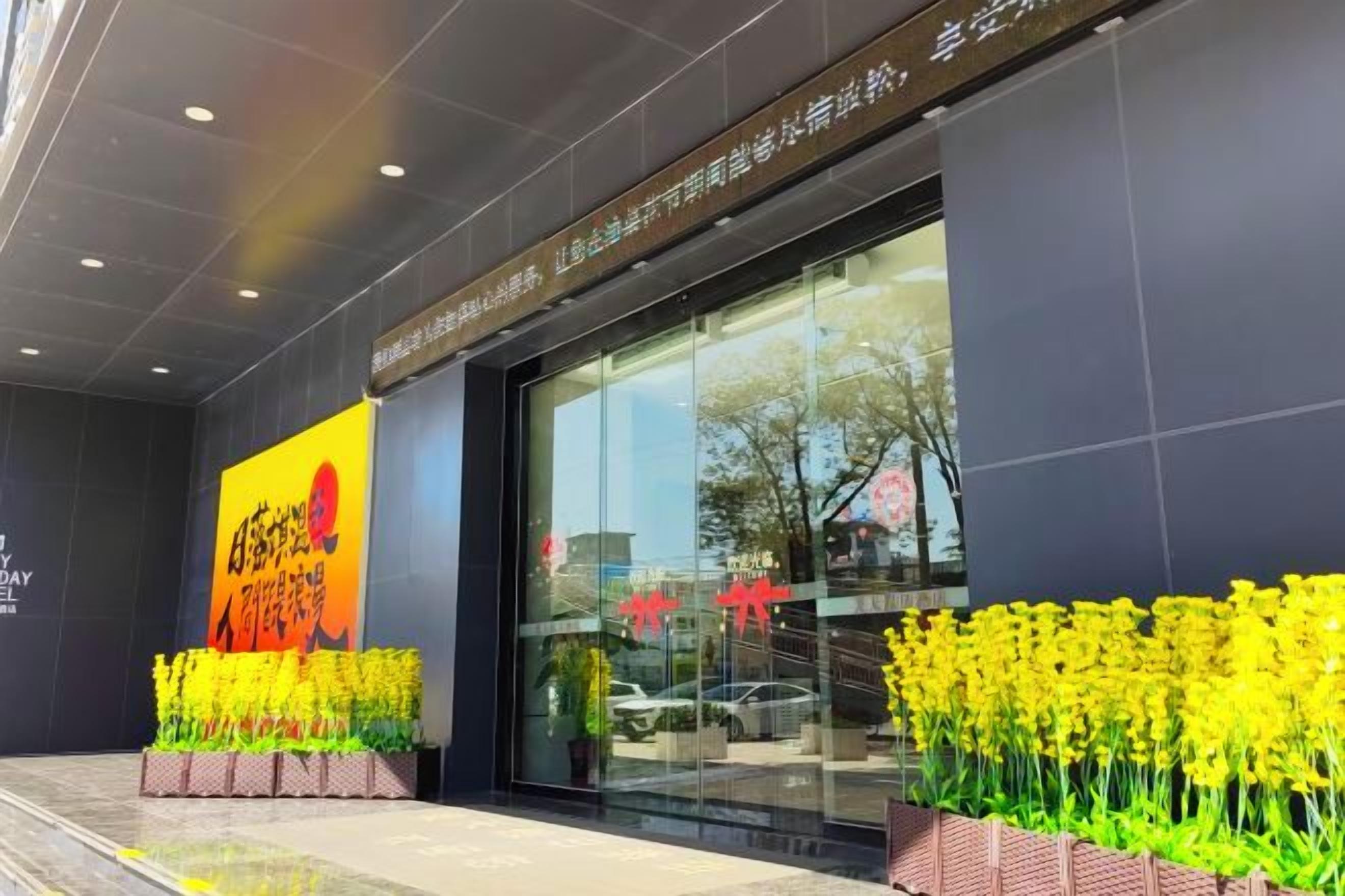A hotel entrance with a large glass facade, a signboard, and yellow flowers in front.