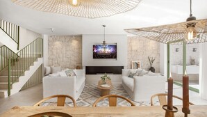 Un salon moderne comprenant un ensemble de canapés, un téléviseur fixé à un mur en pierre, un escalier avec des rampes vertes et un coin repas avec une table en bois.