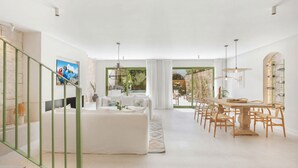 Un salon moderne avec un canapé, une table à manger et des chaises, un escalier avec des rampes vertes, et une vue sur un jardin à travers de grandes fenêtres.